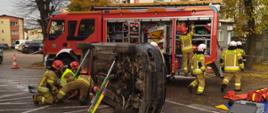 Zdjęcie przedstawia scenę ćwiczeń ratownictwa technicznego straży pożarnej w warunkach miejskich. Centralnym punktem jest samochód osobowy przewrócony na dach, wokół którego pracują strażacy w pełnym rynsztunku, symulując udzielanie pomocy ofiarom wypadku drogowego.