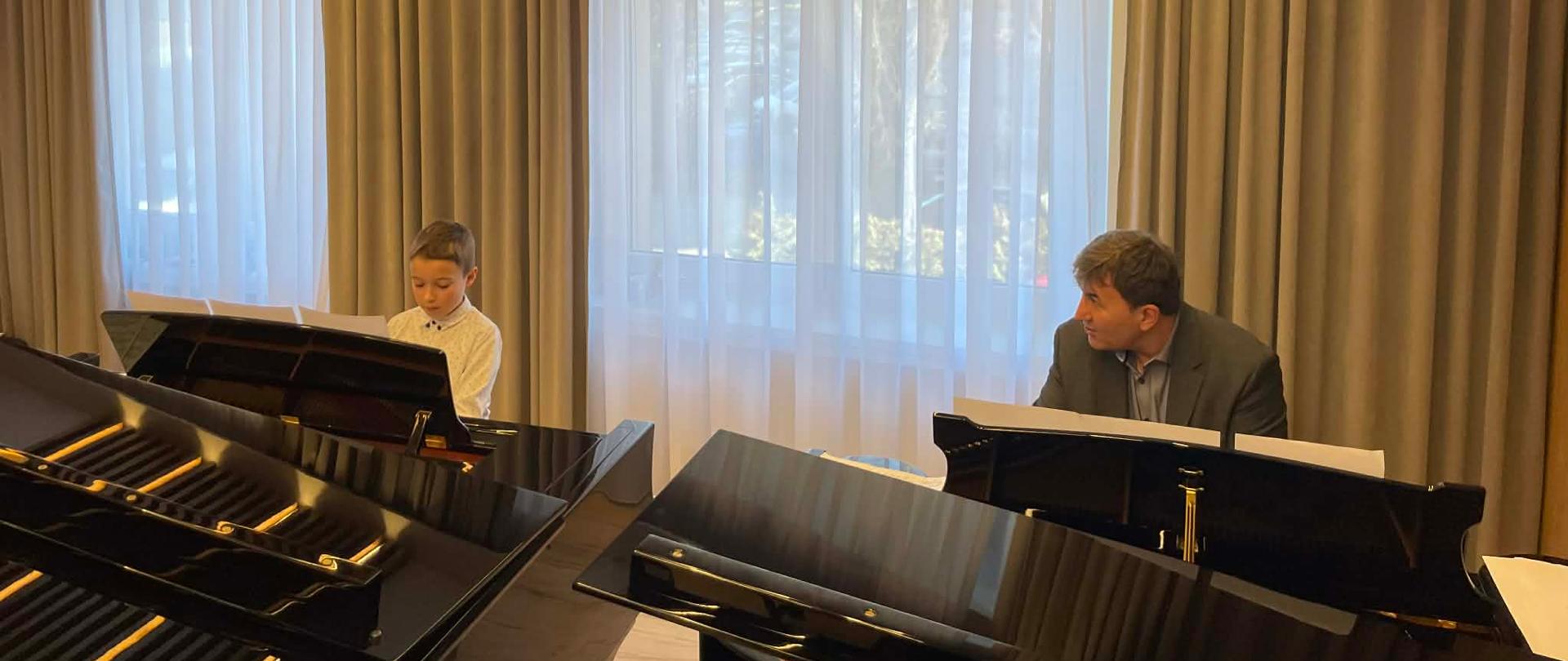 Po lewej stronie przy fortepianie Profesor, przy drugim fortepianie, z prawej strony uczeń. W tle okna.