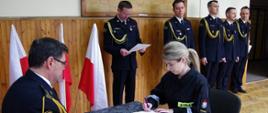 Widok z przodu. Po lewej stronie przy stoliku siedzi komendant powiatowy PSP. Naprzeciwko str. Anna Janeczko w momencie podpisywania dokumentu. Na drugim planie kadra KP PSP w Wadowicach.