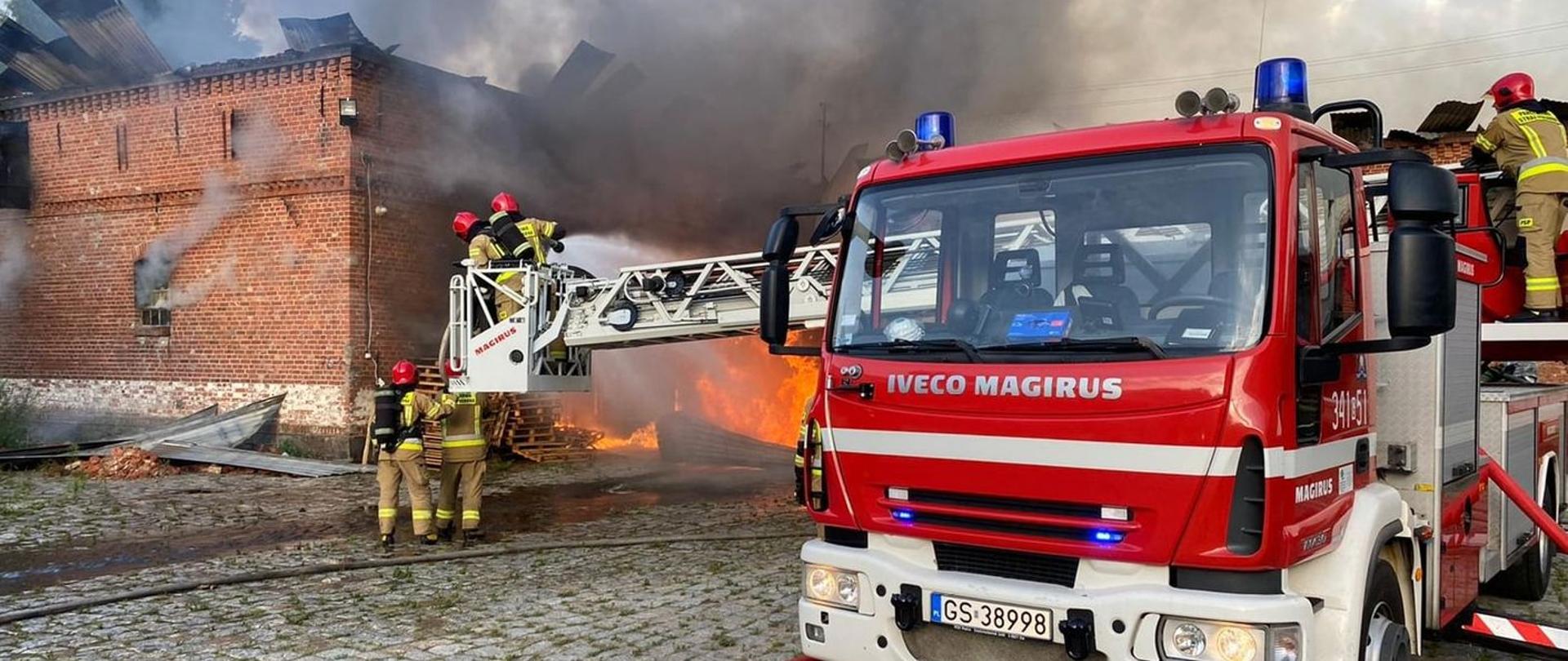 Zdjęcie przedstawia samochód specjalny drabinę wraz z ratownikami gaszącymi płonącą stodołę.