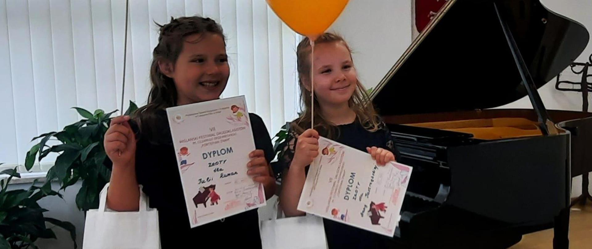 Dwie dziewczynki stoją na scenie przed fortepianem, w rękach trzymają dyplomy, upominki i balony.