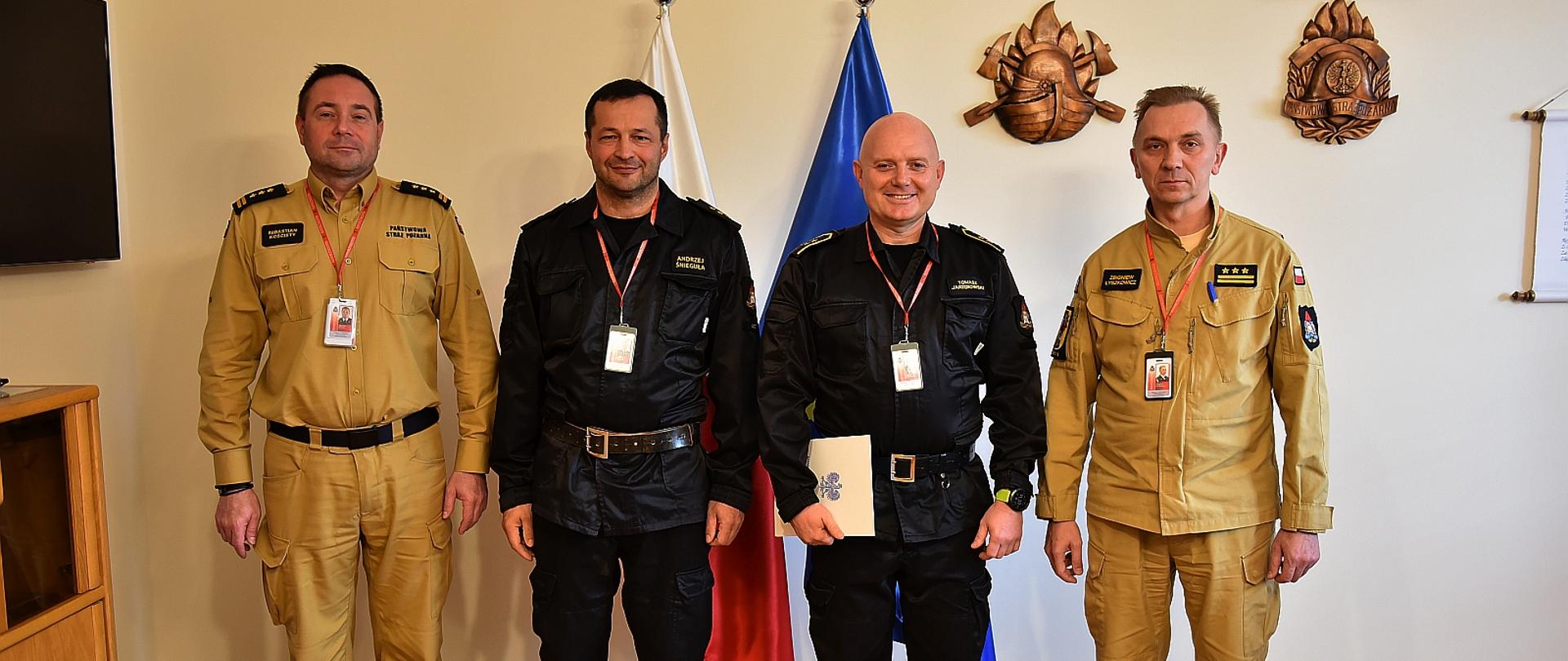 na zdjęciu czterech strażaków w pozujących do zdjęcia na tle ściany za nimi stoją flaga polski i unii europejskiej , strażacy stojący wewnątrz ubrani są w mundurach dowódczo sztabowych dwóch na zewnątrz w mundurach służbowych w kolorze piaskowym