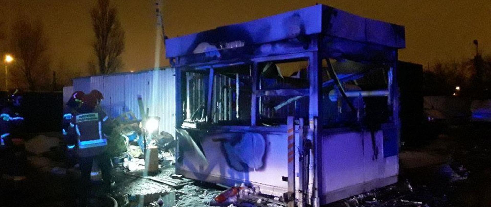 Spalony budynek stróżówki na parkingu, obok trzech strażaków