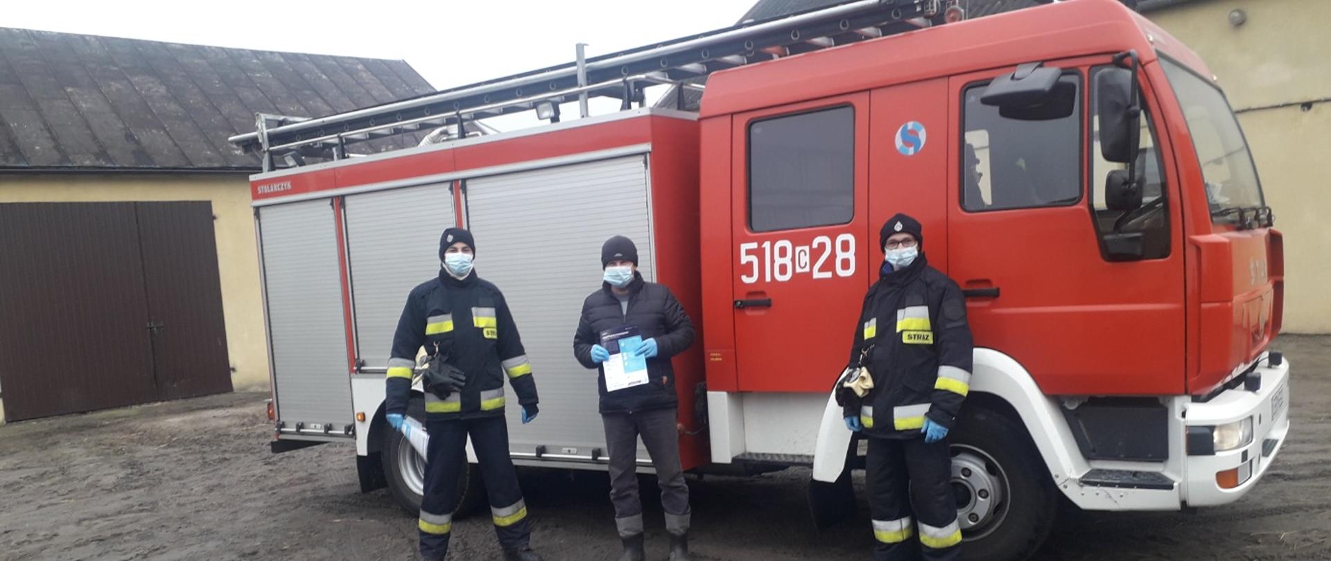 Zdjęcie przedstawia dwóch strażaków OSP, którzy wręczyli mężczyźnie ulotkę dotyczącą szczepień przeciwko COVID-19. Zdjęcie wykonane jest na tle samochodu strażackiego. Na kabinie pojazdu widać numery operacyjne 518c28. Po lewej stronie zdjęcia oraz za samochodem widać budynki gospodarcze. 