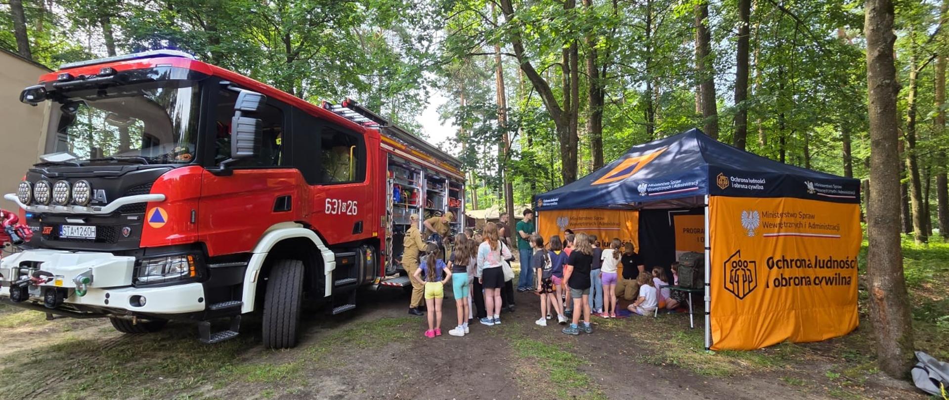 Na zdjęciu widzimy plenerowe spotkanie edukacyjne dla dzieci, zorganizowany na terenie leśnym.
Oto główne elementy widoczne na fotografii:
Wóz strażacki: Po lewej stronie stoi duży, nowoczesny, czerwony samochód ratowniczo-gaśniczy marki Scania (z widocznym oznaczeniem XT na grillu, numerem taktycznym 631[S]26 oraz tablicą rejestracyjną zaczynającą się od STA). Ma otwarte boczne skrytki ze sprzętem ratowniczym, który prezentuje dzieciom strażak w piaskowym mundurze (typu combat).
Namiot promocyjno-edukacyjny: Po prawej stronie stoi granatowo-pomarańczowy namiot ekspozycyjny Ministerstwa Spraw Wewnętrznych i Administracji (MSWiA). Widnieją na nim napisy „Ochrona ludności i obrona cywilna” oraz logo ministerstwa.
Grupa dzieci: Wokół wozu strażackiego oraz pod namiotem gromadzi się spora grupa dzieci w wieku szkolnym. Część z nich przygląda się wyposażeniu wozu, a pozostałe uczestniczą w zajęciach lub prelekcji prowadzonej pod namiotem.
Otoczenie: Całe wydarzenie odbywa się na nieutwardzonym, piaszczystym terenie w otoczeniu wysokich, gęstych drzew, co sugeruje np. teren obozu, lasu lub parku miejskiego.