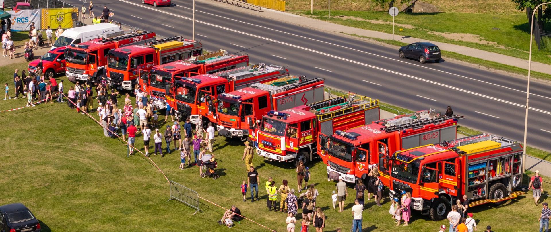 Rząd czerwonych wozów strażackich zaparkowanych na trawiastym placu wzdłuż wielopasmowej drogi podczas festynu plenerowego. Wokół pojazdów gromadzą się dorośli i dzieci.