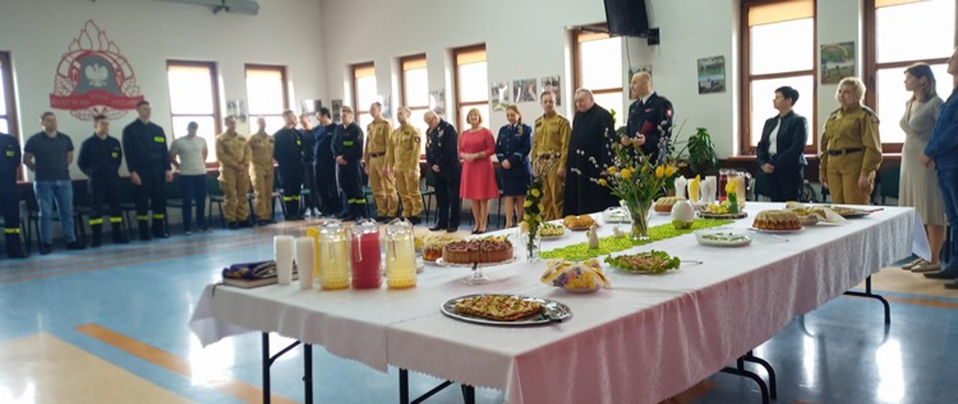 Zdjęcie przedstawia wnętrze dużej, przestronnej sali szkoleniowej w Komendzie Powiatowej w Sokołowie Podlaskim. Na pierwszym planie widoczny jest długi stół przykryty białym obrusem, na którym ustawiono różnorodne potrawy i napoje, w tym ciasta, sałatki, owoce i dzbanki z sokiem. Stół jest ozdobiony wiosennymi elementami, takimi jak gałązki z zielonymi listkami i małe żółte ozdoby, co może wskazywać na świąteczne lub okolicznościowe spotkanie.
W tle, wzdłuż ściany po lewej stronie, stoi rząd mężczyzn w różnych mundurach strażackich – zarówno w ciemnych, jak i jasnobeżowych. Po prawej stronie stołu, również wzdłuż ściany, stoi mniejsza grupa osób, wśród których są zarówno mężczyźni w mundurach strażackich, jak i kobiety oraz mężczyźni w ubraniach cywilnych. Na ścianie po lewej stronie widnieje emblemat lub godło Państwowej Straży Pożarnej. Sala jest jasna, z dużymi oknami widocznymi w tle, a podłoga ma geometryczny wzór w odcieniach szarości i brązu.