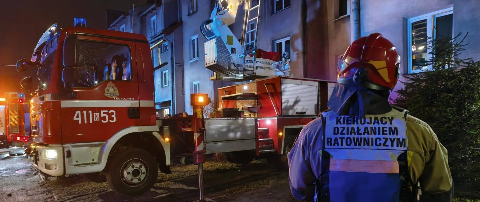 Zdjęcie zrobione nocą. Przy budynku mieszkalnym stoi rozstawiony podnośnik hydrauliczny staży pożarnej. Z prawej strony zdjęcia stoi strażak w czerwonym hełmie i kamizelce z napisem kierujący działaniem ratowniczym.