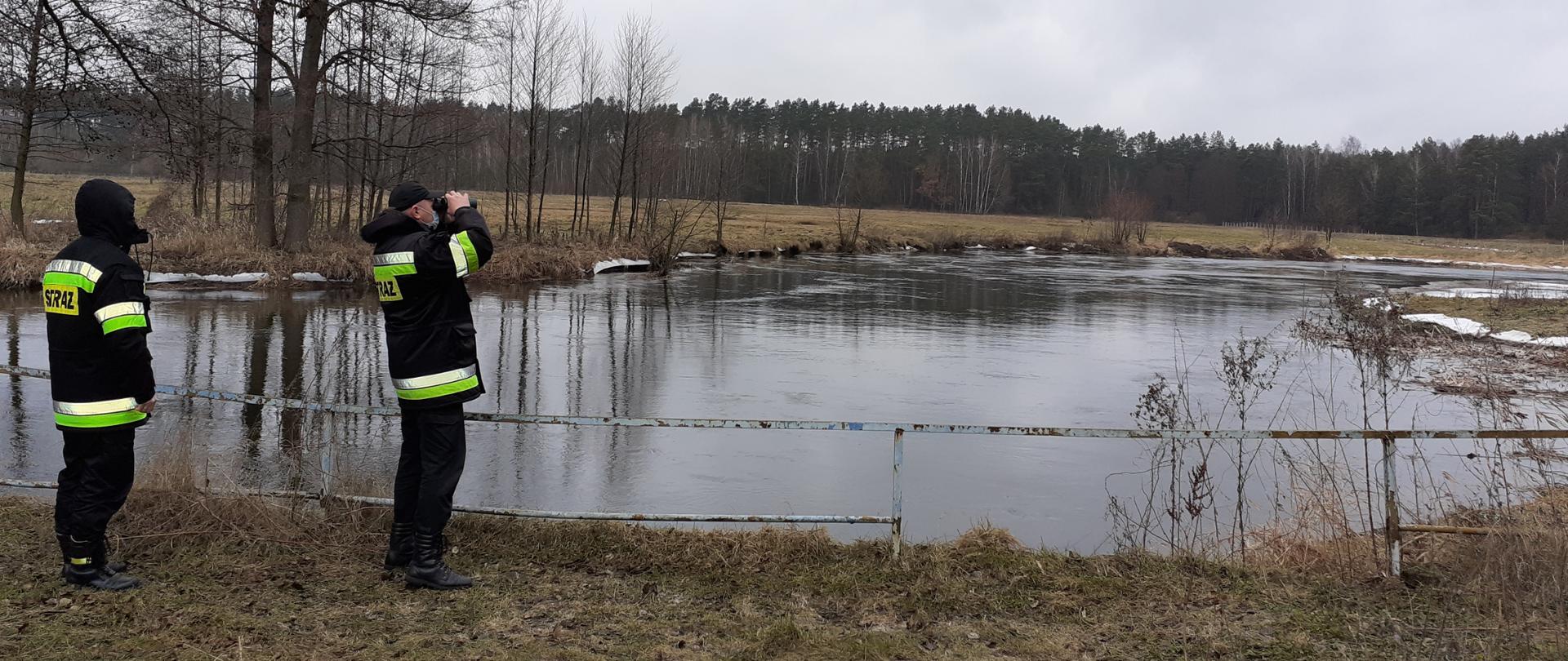 Na pierwszym planie widać rzekę oraz dwóch strażaków obserwujących stan wody. Jeden z nich patrzy przez lornetkę.