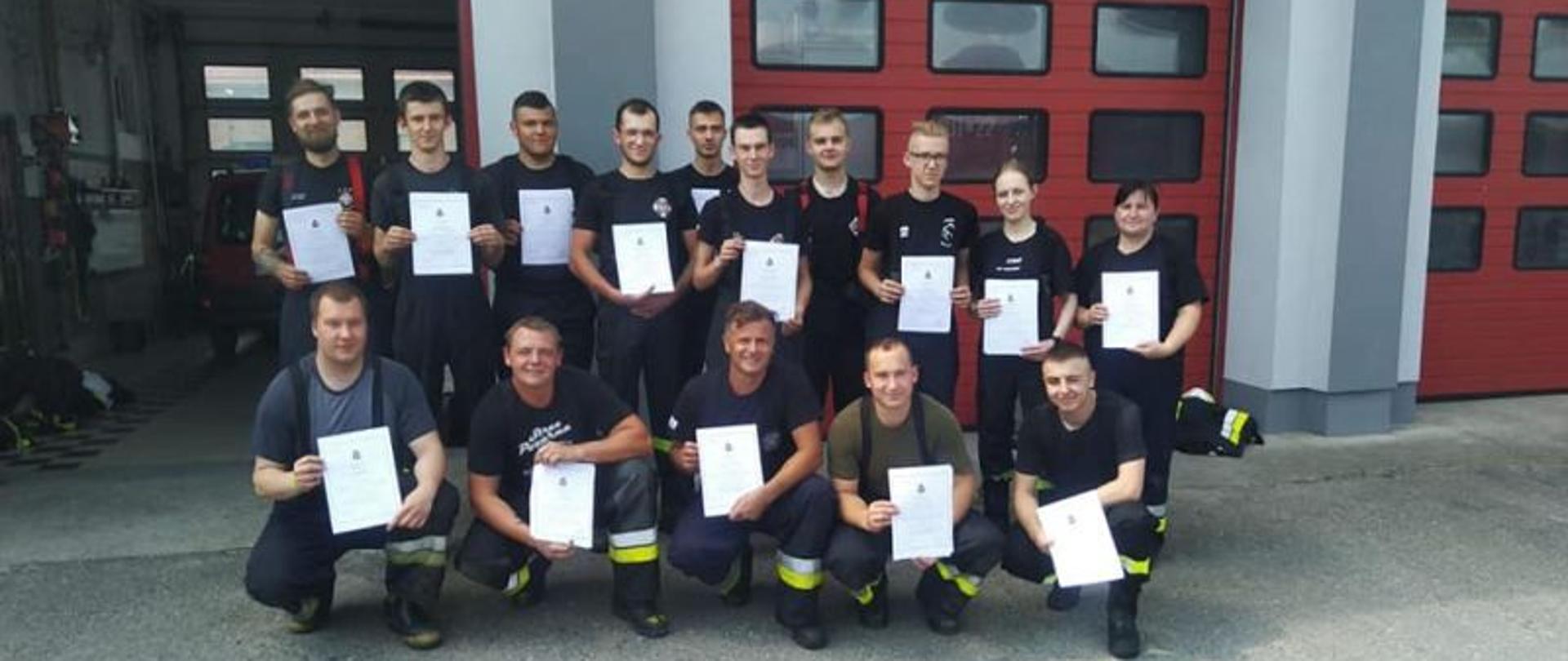 Zdjęcie grupowe. Na zdjęciu wszyscy uczestnicy kursu z dyplomami.Strażacy i strażaczki stoją w dwuszeregu. Pierwszy szereg kuca. W tle garaże KP PSP Białogard.