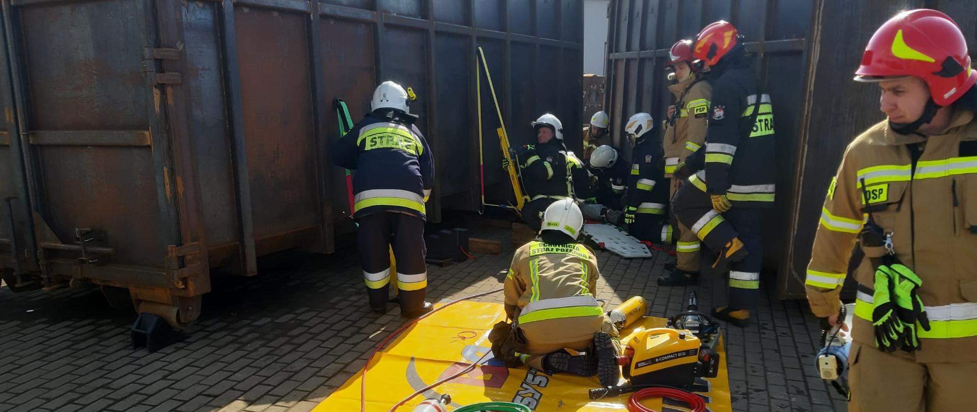 Na zdjęciach widać strażaków podczas ćwiczeń, w tle metalowe kontenery, n pierwszym planie rozłożona żółta mata ze sprzętem hydraulicznym
