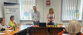 Kobieta i mężczyzna stojąc przemawiają na sali konferencyjnej. Obok przy stolikach siedzą inne kobiety. Na stole stoją komputer, szklanki, woda w butelkach oraz ciasto i cukierki. W tle baner reklamowy oraz godło Polski