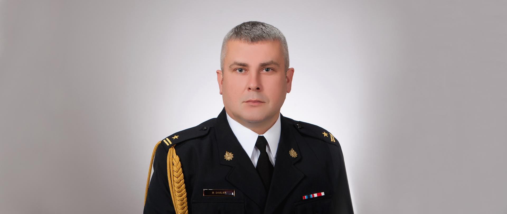 Zdjęcie na szarym tle Zastępcy Komendanta Powiatowego PSP w Nysie mł. bryg. Marcina Skalnego w mundurze galowym. 