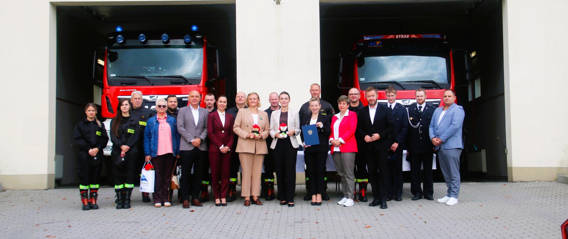 grupa osób stoi przed remizą, w które stoją dwa ciężkie samochody ratowniczo gaśnicze