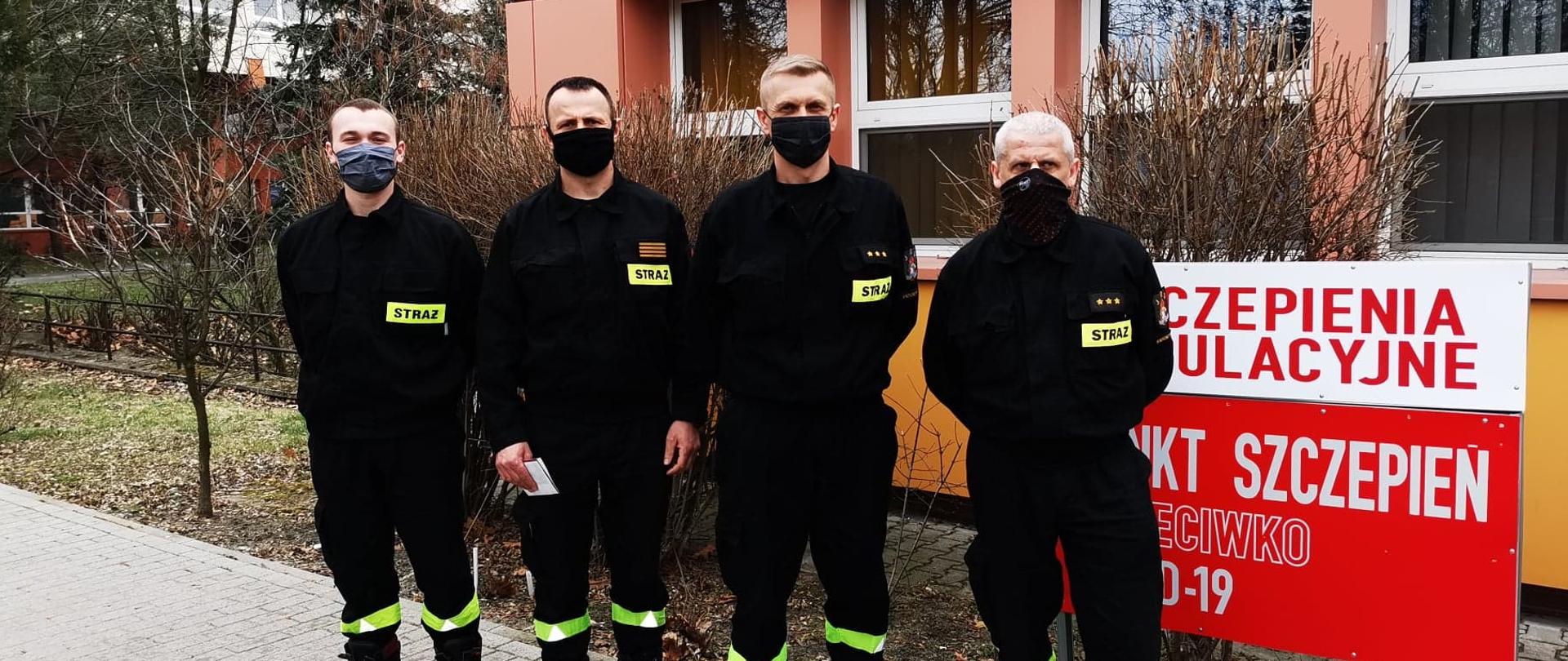 Czterech funkcjonariuszy KP PSP w Krotoszynie w ubraniach koszarowych stoi w szeregu przed punktem szczepień. Zdjęcie po odbytych szczepieniach przeciw COVID-19.