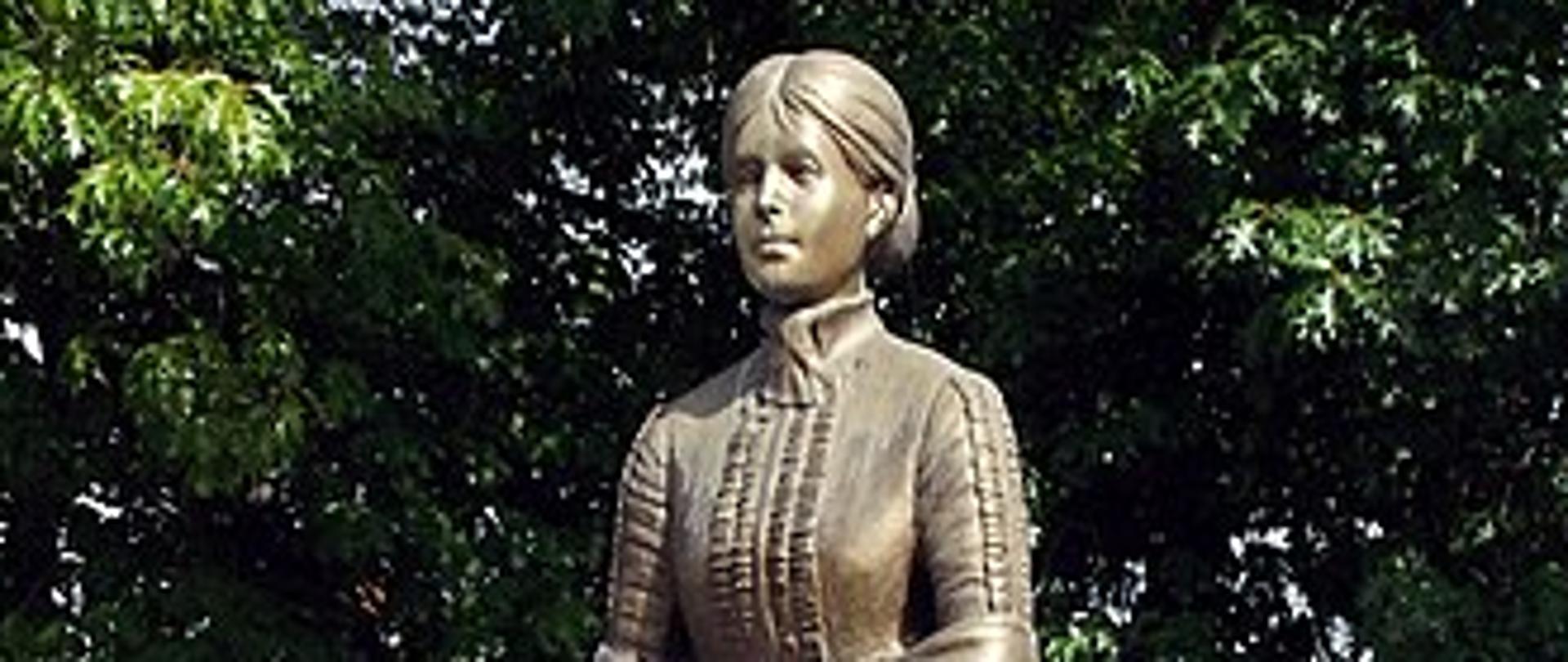 zdjęcie pomnika Marii Konopnickiej z dzieckiem, kolor brązowy, na tle drzew i w oddali budynków