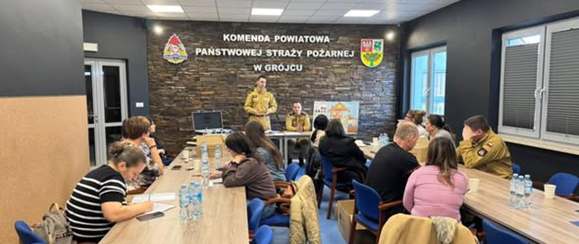 Szerszy widok sali konferencyjnej. Na ścianie z tyłu znajduje się napis wskazujący, że to Komenda Powiatowa Państwowej Straży Pożarnej w Grójcu. Na środku siedzi dwóch strażaków przy stole, jeden z nich prowadzi spotkanie. Uczestnicy siedzą po obu stronach stołów, część notuje, inni słuchają. Na stołach stoją butelki z wodą i leżą materiały szkoleniowe. Pomieszczenie jest dobrze oświetlone, z ciemnymi ścianami i jasnym sufitem.