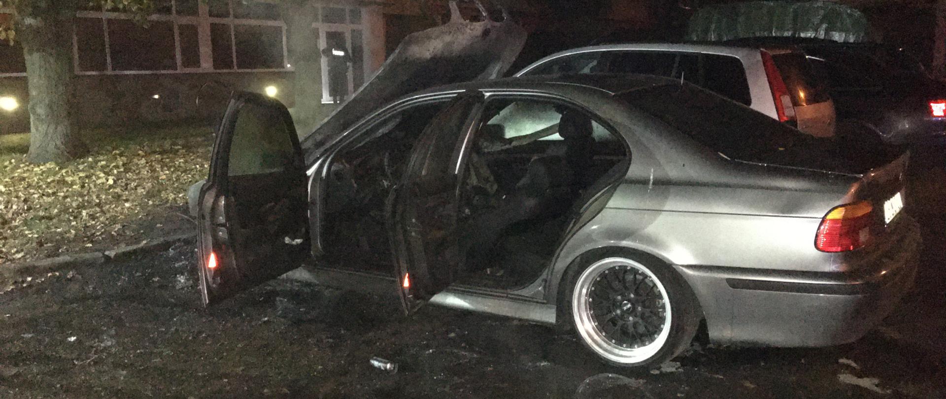 Na zdjęciu widnieje częściowo spalony srebrny pojazd. Zdjęcie zostało wykonane od strony jego lewego boku. Drzwi pojazdu są otwarte, a maska pojazdu zasłania przednią szybę. Środek pojazdu częściowo spalony. Za pojazdem zaparkowane na parkingu są następne pojazdy. Po lewej stronie zdjęcia znajduje się drzewo, a za nim znajduje się blok mieszkalny.