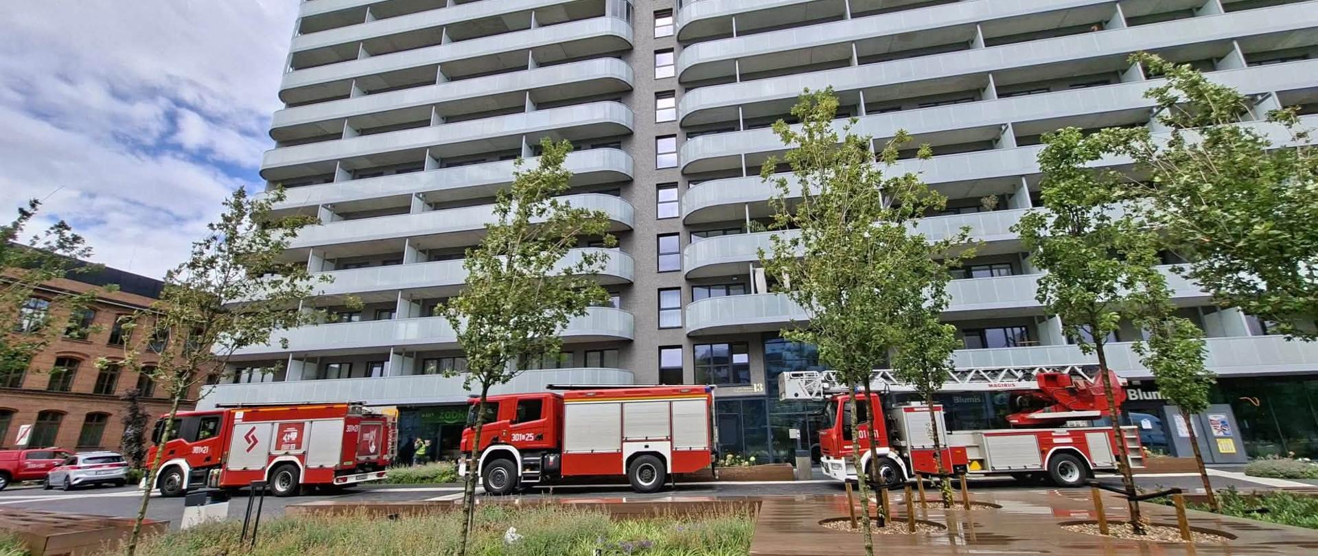 Trzy czerwone wozy strażackie zaparkowane przed nowoczesnym, wysokim wieżowcem mieszkalnym z białymi balkonami. Na pierwszym planie widać zielony skwer z młodymi drzewkami.
