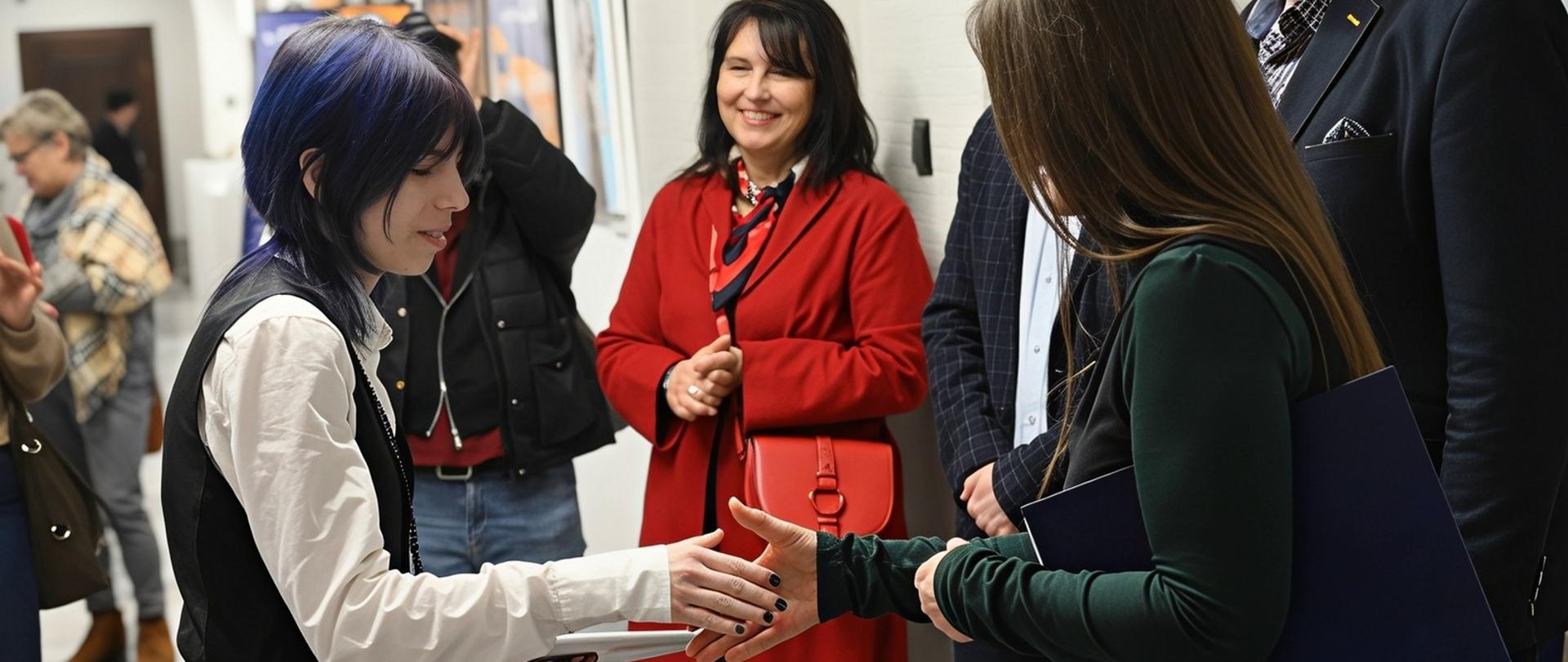 Kobieta w długich włosach gratuluje Laureatce konkursu podając jej dłoń, Wokół stoją uśmiechnięci ludzie; na ścianach galerii wpiszą prace konkursowe