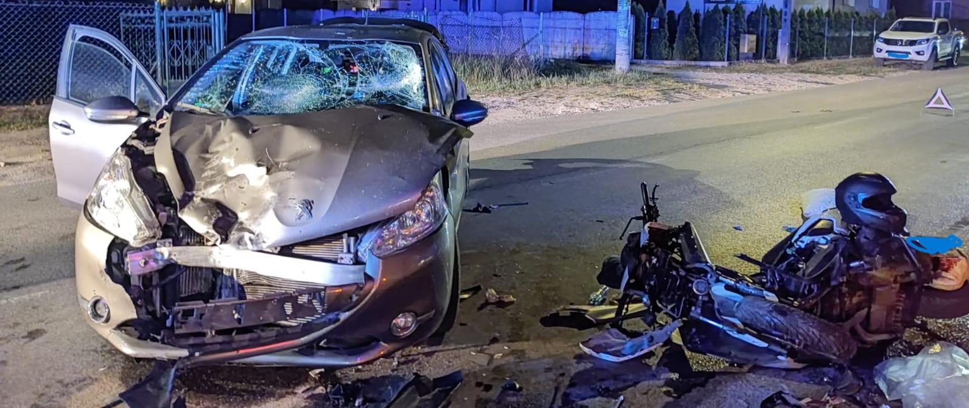 Na zdjęciu dwa pojazdy po zderzeniu znajdujące się na drodze asfaltowej. W dolnej części zdjęcia widoczny jest leżący na boku motocykl z poważnymi uszkodzeniami w przedniej części, na jednośladzie leży kask. Za nim znajduje się samochód osobowy ze zniszczoną maską i zderzakiem oraz pękniętą szybą czołową. Wokół pojazdów porozrzucane są plastiki oraz wysypany sorbent na plamy oleju. Pojazdy znajdują się na drodze a w tle widoczne są budynki mieszkalne.
