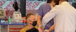 Zdjęcie przedstawia komendanta głównego psp nadbrygadiera Andrzeja Bartkowiaka podczas zabiegu szczepienia szczepionką przeciw COVID-19, obok niego stoją pracownicy służby zdrowia, w tle szpitalu tymczasowym na PGE Narodowym.