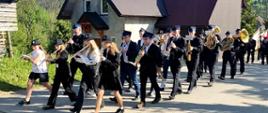 Pochód orkiestry ulicami Jaworek. Orkiestra ubrana w wizytowe stroje strażackie. W tle dom. Wszyscy członkowie graja na instrumentach dętych. 