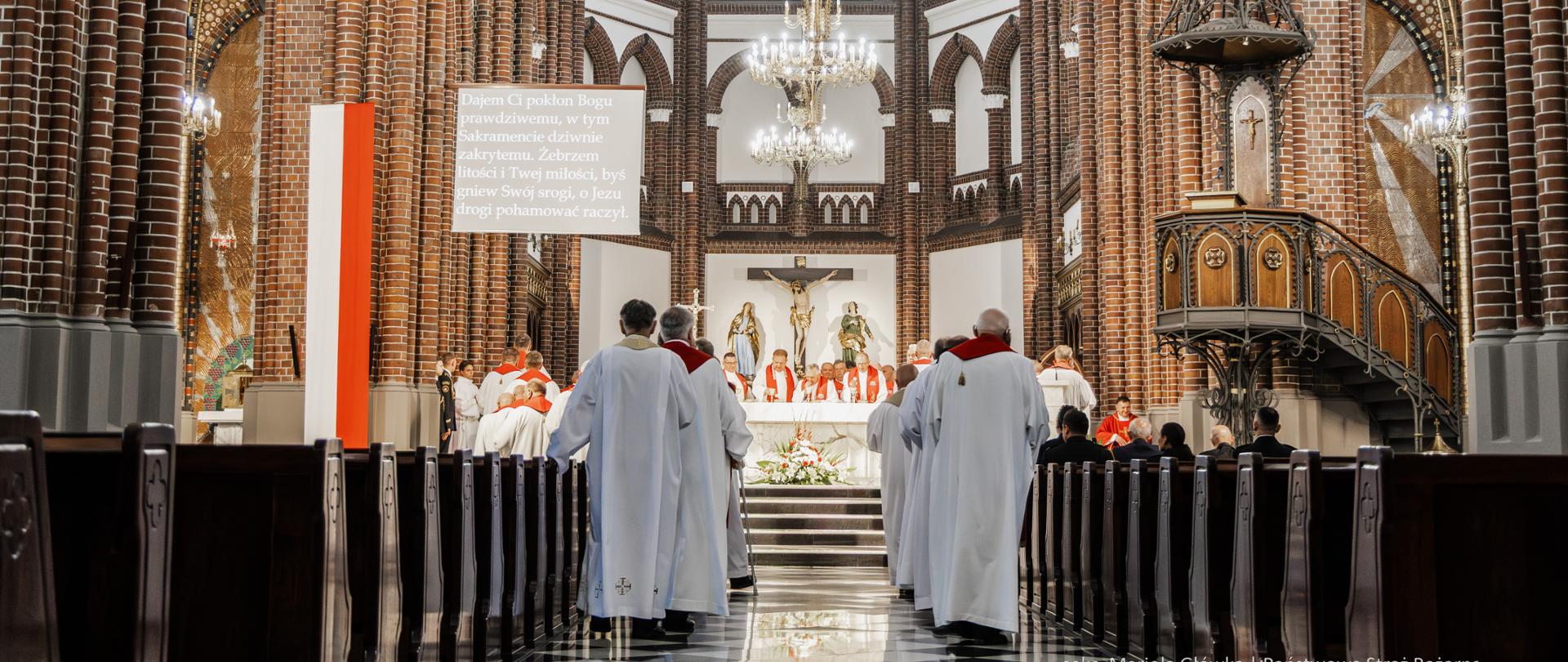 Widok wnętrza kościoła podczas uroczystej mszy. Przy ołtarzu zgromadzeni są duchowni w szatach liturgicznych, a nawą w ich kierunku idą księża w białych albach. Wnętrze świątyni zdobią ceglane kolumny, witraże i żyrandole.