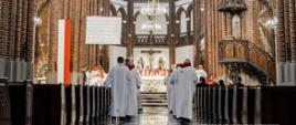 Widok wnętrza kościoła podczas uroczystej mszy. Przy ołtarzu zgromadzeni są duchowni w szatach liturgicznych, a nawą w ich kierunku idą księża w białych albach. Wnętrze świątyni zdobią ceglane kolumny, witraże i żyrandole.