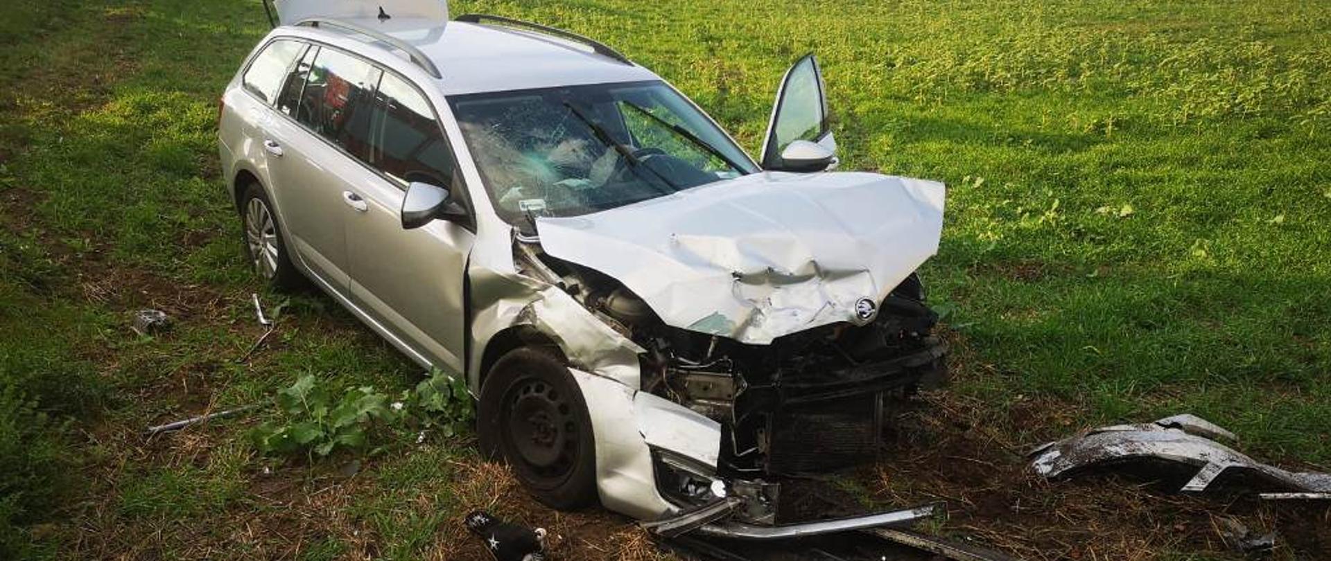 Srebrny samochód osobowy kombi z uszkodzonym przodem, stojący na zielonej łące. W około porozrzucane elementy karoserii. W oddali widać stojących ludzi.