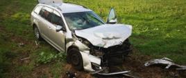 Srebrny samochód osobowy kombi z uszkodzonym przodem, stojący na zielonej łące. W około porozrzucane elementy karoserii. W oddali widać stojących ludzi.