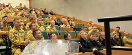 Sala szkoleniowa. W rzędach na krzesłach siedzą strażacy w umundurowaniu w kolorze żółtym i czarnym.
