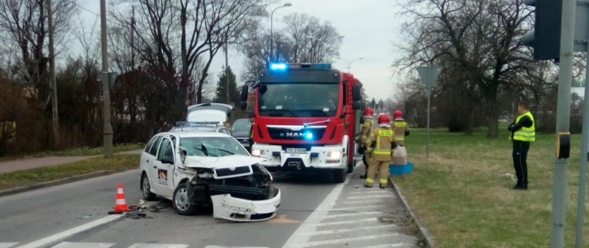 Zdjęcie przedstawia samochód osobowy po zderzeniu. Na dalszym planie samochód pożarniczy oraz grupa strażaków.