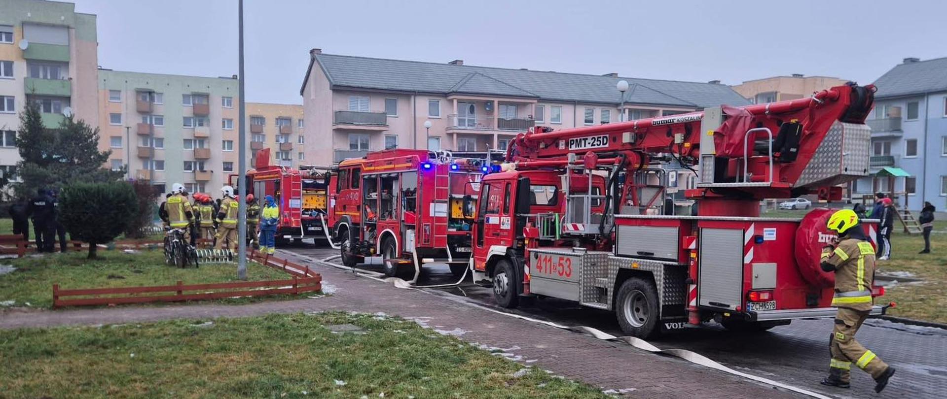 Osiedle bloków mieszkalnych. Na pierwszym planie widać trzy pojazdy straży pozarnej w tym jeden podnośnik hydrauliczny zaparkowane na jezdni. Przy nich grupa strażaków. W tle znajdują się bloki mieszkalne. 