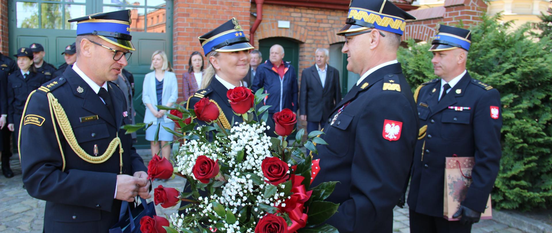 Komendant generał przyjmuje kwiaty od pracowników. Bukiet czerwonych róż. W tle fragment budynku komendy, współpracownicy.