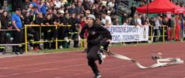 Młoda strażaczka dynamicznie biegnie po czerwonej torze bieżni lekkoatletycznej z prądownicą w ręku i ciągniętym wężem pożarniczym podczas konkurencji rozwinięcia bojowego na zawodach sportowo-pożarniczych.