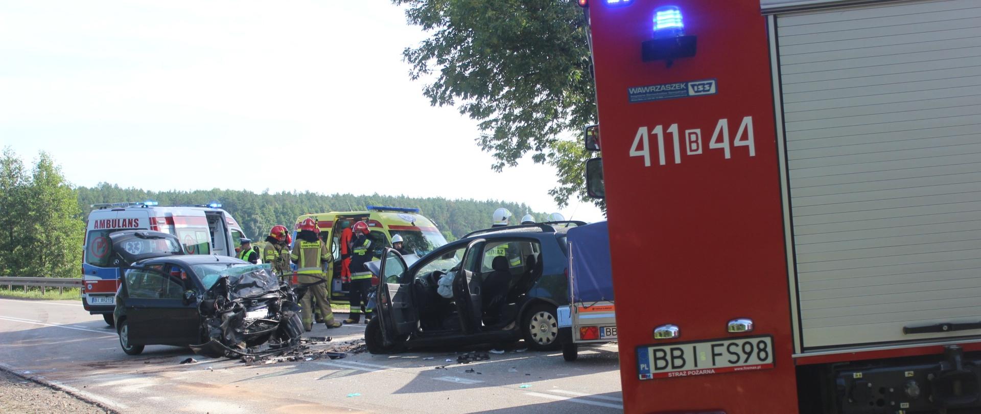 Rozbity samochód osobowy po czołowym zderzeniu. W tle działający na miejscu zdarzenia strażacy oraz 2 karetki pogotowia ratunkowego. Z boku widoczny samochód ratowniczo-gaśniczy.