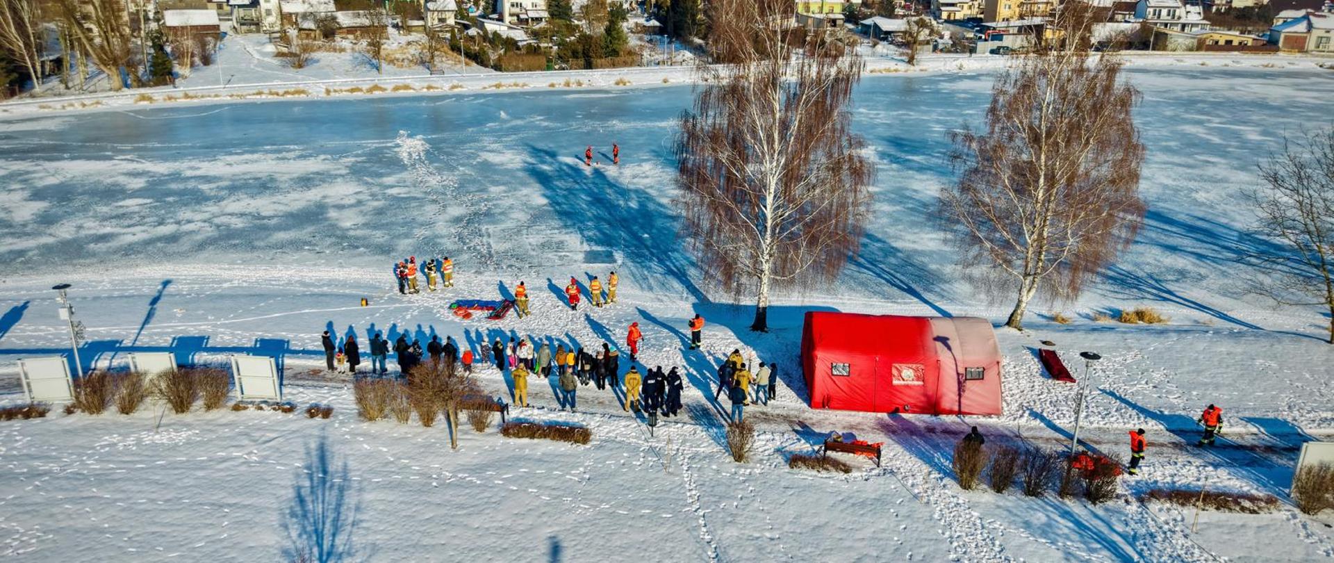 Widok z drona na zamarznięty, pokryty śniegiem zbiornik wodny w Lipsku zimą. Na lodzie oraz na brzegu widoczna jest duża grupa ludzi, w tym ratownicy w pomarańczowych i czerwonych strojach oraz strażacy. Na brzegu rozstawiony jest duży, czerwony namiot pneumatyczny. Ratownicy na lodzie prezentują sprzęt ratunkowy (m.in. sanie lodowe). W tle widać panoramę domów jednorodzinnych oraz bezchmurne, błękitne niebo. Długie cienie sugerują mroźny, słoneczny dzień.