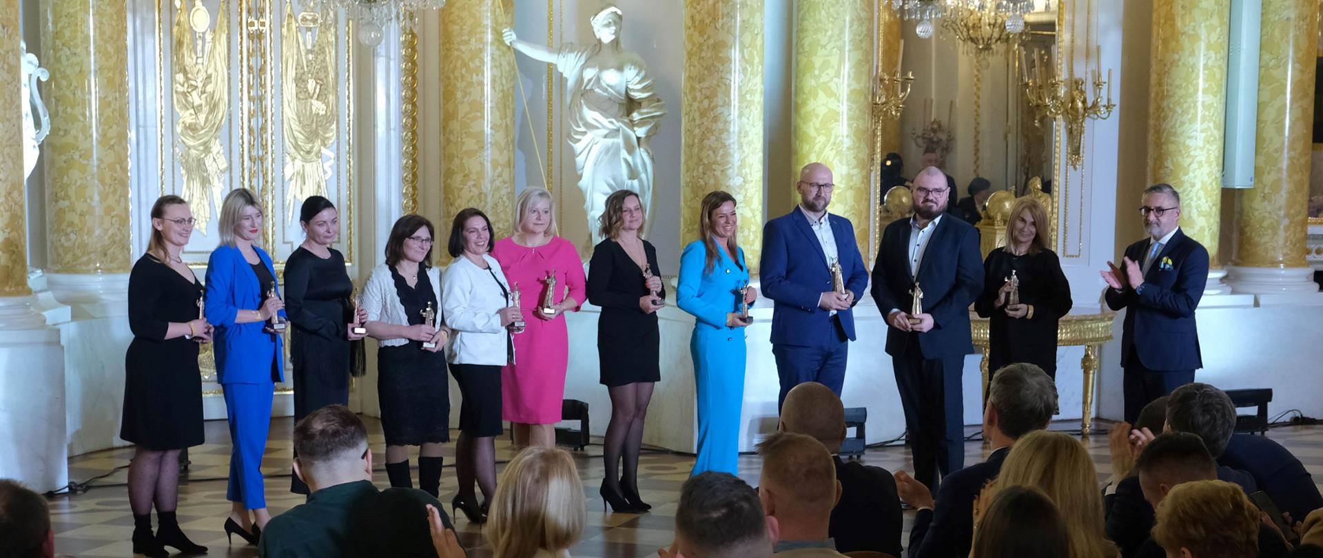 Grupa kobiet i mężczyzn stoi w rzędzie ze statuetkami w rękach. Przed nimi osoby siedzą na krzesłach i robią zdjęcia.