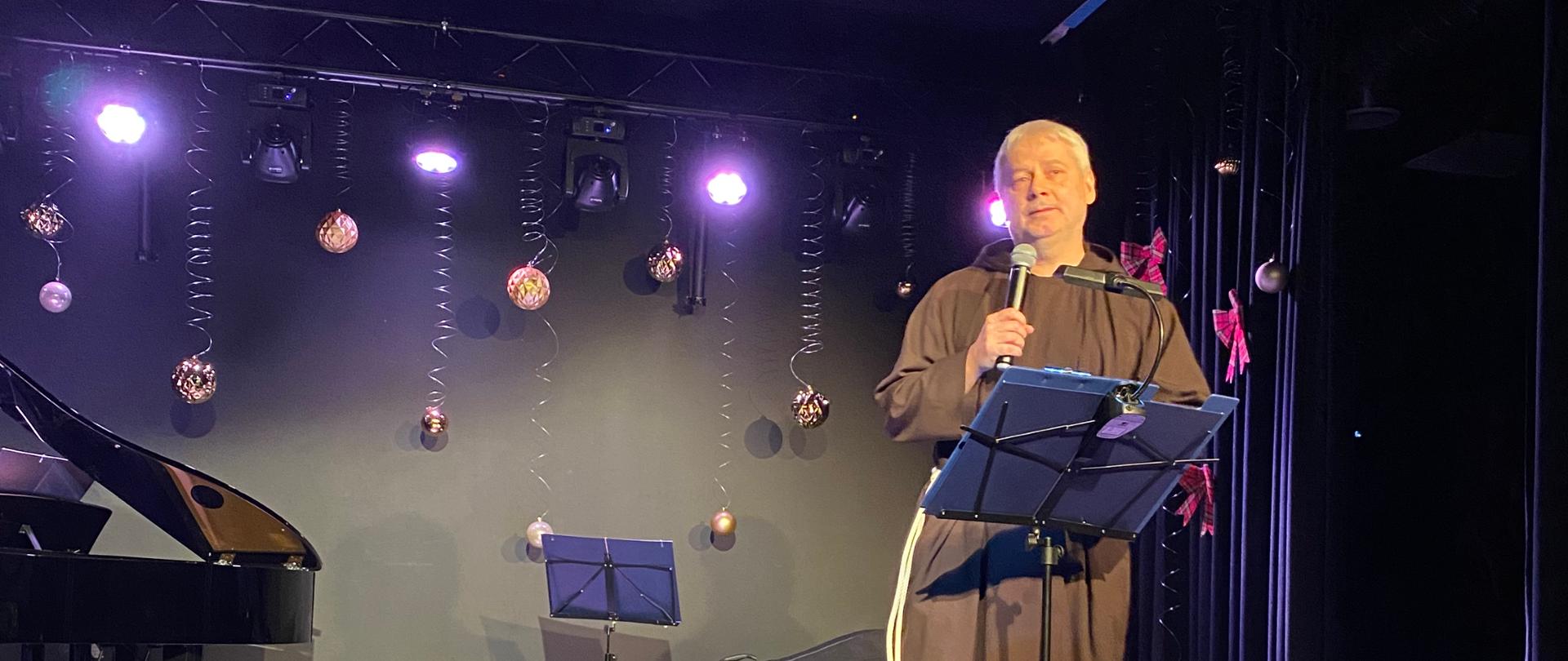 Na zdjęciu widać mężczyznę stojącego na scenie, oświetlonej fioletowymi i ciepłymi światłami. Ma na sobie brązowy habit z jasnym sznurem przewiązanym w pasie. Stoi wyprostowany przy mikrofonie umieszczonym na statywie, przed nim znajduje się pulpit. Tło sceny jest ciemne, udekorowane wiszącymi bombkami i delikatnymi spiralnymi ozdobami. Po lewej stronie sceny stoi fortepian z uniesioną klapą, a obok widoczny jest stojak na nuty oraz futerał na instrument muzyczny leżący na podłodze. Na podłodze, przy krawędzi sceny, ustawiono lampiony ze świecami. Przed sceną znajduje się mały stolik przykryty białym obrusem, na którym leży stos kartek lub kopert ułożonych w formie wachlarza.
