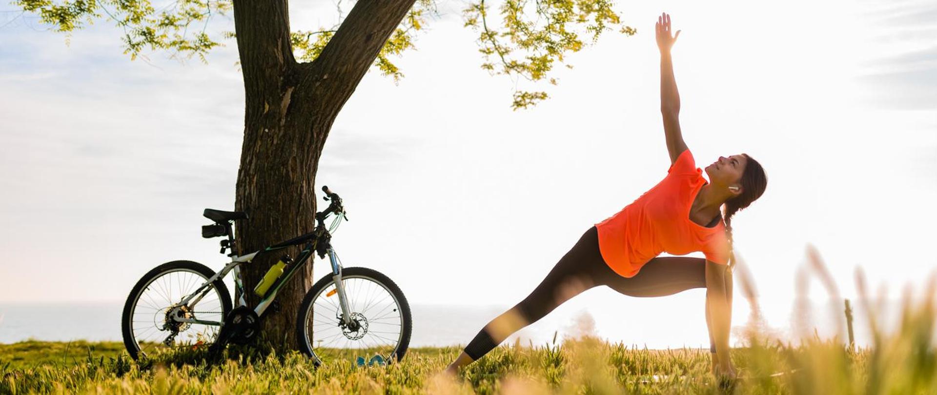 Szczupła kobieta w kolorowym stroju fitness uprawiająca jogę na świeżym powietrzu. Po lewej stronie w tle rower oparty o drzewo.