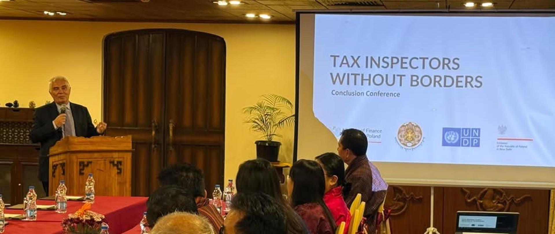 Dr Piotr Antoni Świtalski przemawia podczas konferencji podsumowującej projekt Tax Inspectors Without Borders w Bhutanie. Przedstawiciele bhutańskich instytucji i partnerów projektu siedzą przy stołach konferencyjnych, a na ekranie widoczna jest nazwa programu.