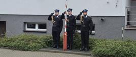 Trzech strażaków w ubraniach wyjściowych podczas uroczystego śpiewania hymnu państwowego na tle budynku komendy 