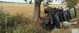Zdjęcie przedstawia samochód osobowy leżący na boku w przydrożnym rowie obok drzewa. Samochód całkowicie zniszczony.