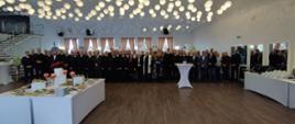 Na zdjęciu widoczna jest liczna grupa uczestników spotkania wigilijnego zgromadzona w przestronnej sali. W pierwszym planie znajdują się stoły z przygotowanymi potrawami wigilijnymi oraz dekoracjami świątecznymi. W tle ustawieni są funkcjonariusze PSP, zaproszeni goście oraz przedstawiciele duchowieństwa. Uczestnicy stoją w uporządkowanym szyku, co podkreśla uroczysty charakter wydarzenia. Całość dopełnia elegancki wystrój sali i świąteczna iluminacja sufitowa.