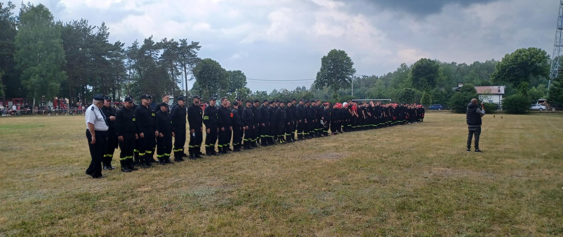 Druhny i druhowie stoją w dwuszeregu na stadionie podczas otwarcia zawodów sportowo-pożarniczych Ochotniczych Straży Pożarnych powiatu człuchowskiego