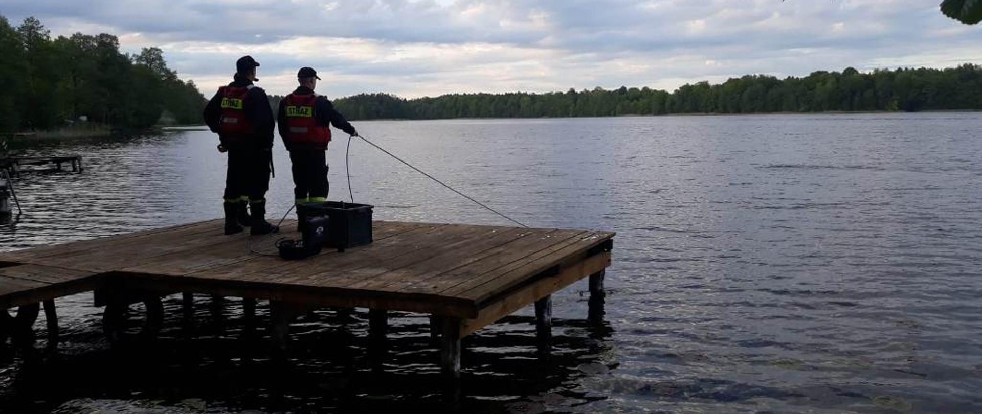 Widok dwóch strażaków na pomoście zabezpieczających strażaka-nurka poszukującego zatopionej łodzi.