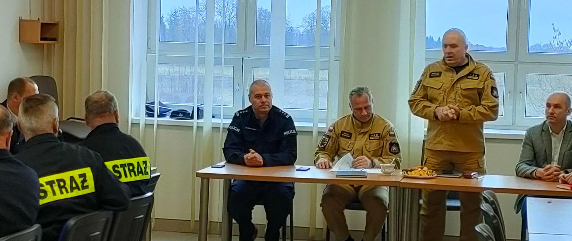 Kilkunastu strażaków siedzi przy stołach w świetlicy. Na wprost za stołem prezydialnym siedzą cztery osoby. Na stojąco przemawia Komendant Powiatowy PSP w Rypinie starszy brygadier Grzegorz Łydkowski.