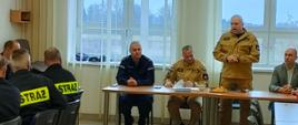 Kilkunastu strażaków siedzi przy stołach w świetlicy. Na wprost za stołem prezydialnym siedzą cztery osoby. Na stojąco przemawia Komendant Powiatowy PSP w Rypinie starszy brygadier Grzegorz Łydkowski.