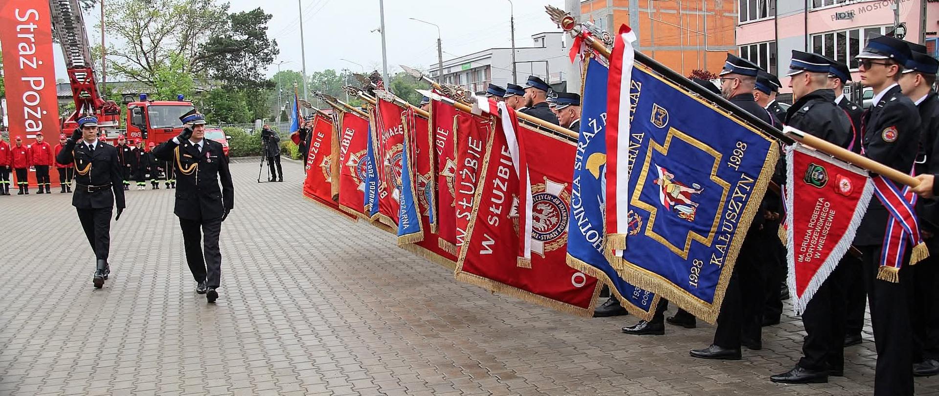 Na zdjęciu widać uroczystość strażacką odbywającą się na placu przed budynkiem (prawdopodobnie remizą strażacką).
Po prawej stronie stoi szpaler strażaków w galowych mundurach – mają czarne czapki z daszkiem i trzymają liczne sztandary w kolorach czerwonym i niebieskim, ozdobione złotymi haftami oraz frędzlami. Sztandary są ustawione pod kątem, tworząc jednolitą, reprezentacyjną linię.
Po lewej stronie dwóch strażaków maszeruje wzdłuż tego szpaleru – jeden z nich oddaje salut, co wskazuje na oficjalny charakter wydarzenia (np. przegląd pododdziałów lub ceremonia).
W tle widać:
czerwone wozy strażackie,
wysuniętą drabinę pożarniczą,
grupę innych strażaków ustawionych w szeregu,
budynki oraz drzewa.
Na banerze po lewej stronie widnieje napis związany ze strażą pożarną (w języku polskim), co sugeruje, że zdjęcie zostało wykonane w Polsce.
Całość przedstawia podniosłą, ceremonialną atmosferę – prawdopodobnie święto strażaków, przekazanie sztandaru lub inna oficjalna uroczystość